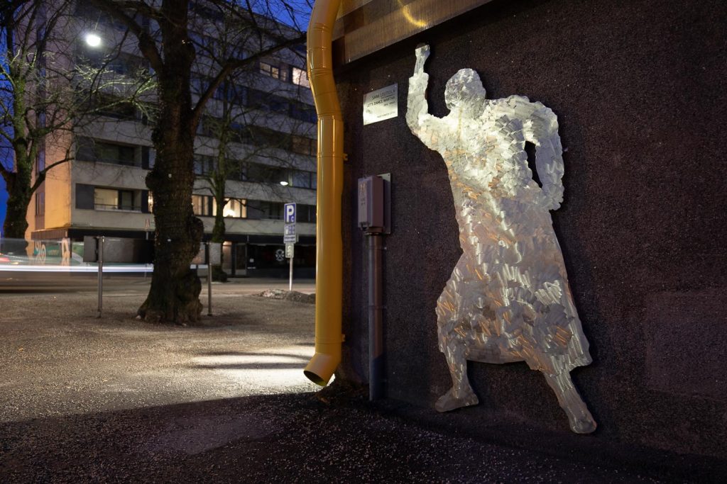 Seinään kiinnitetty metallinen ihmishahmoa esittävä veistos pimenevässä illassa.