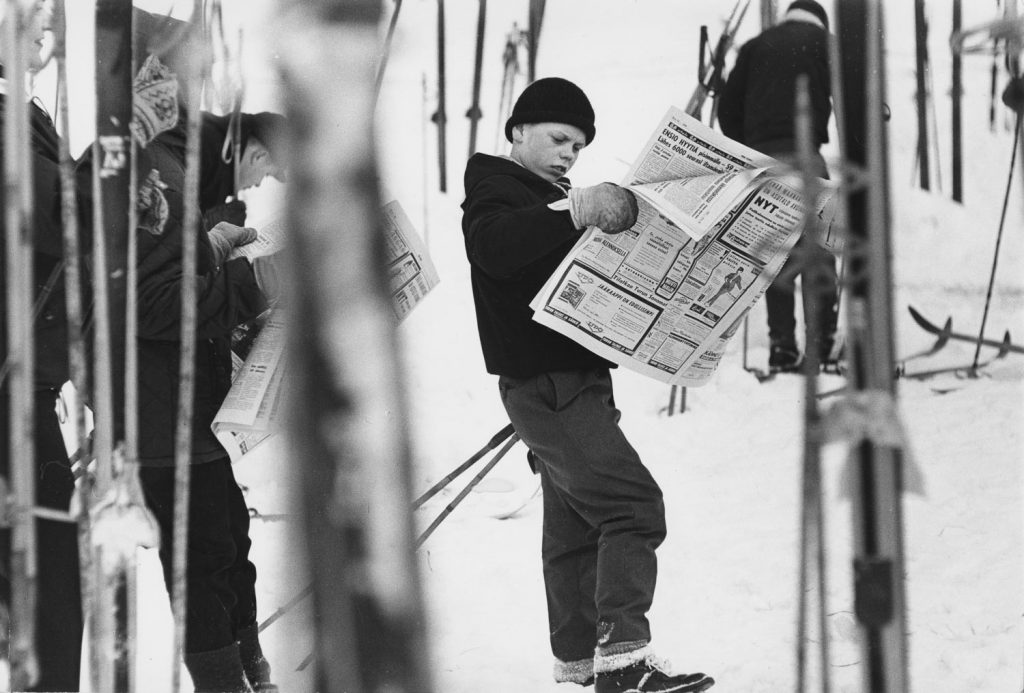 Mustavalkoisessa kuvassa poika lukee sanomalehteä lumihankeen pystyn laitettujen suksien keskellä.