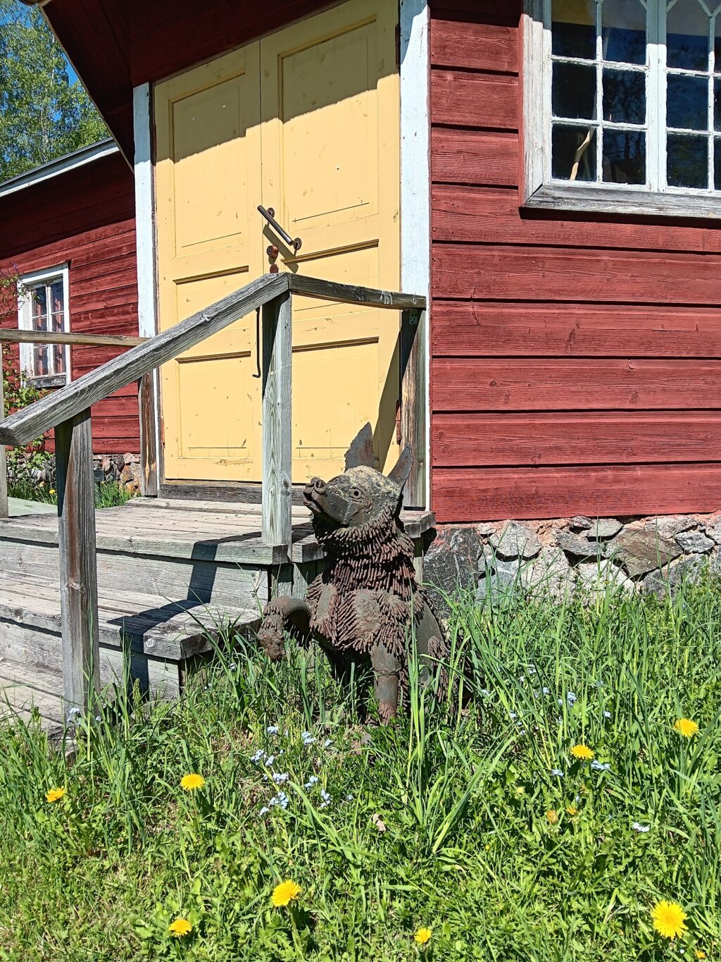 Metallinen koiraveistos punaiseksi maalatun, vanhan puurakennuksen puisten portaiden vieressä.
