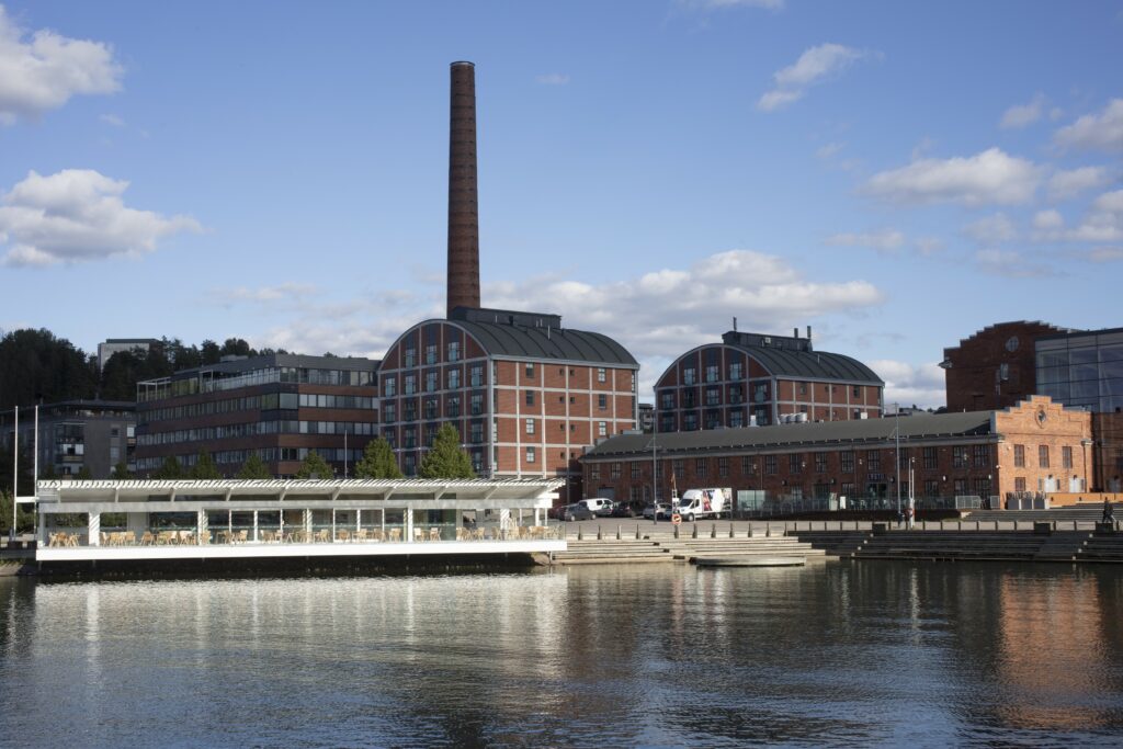 Punatiilisiä rakennuksia rannassa, rakennusten takana kohoaa piippu, taivas on sininen.