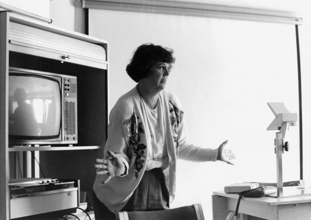 Nainen elehtii käsillään opettaessaan ja taustalla televisio ja piirtoheittimen valkokangas.