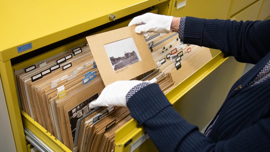 Museotyöntekijä tutkii mustavalkoista valokuvaa, jonka hän on ottanut keltaisesta arkistokaapista.