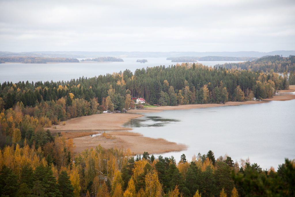 Pilvisenä päivänä korkealta kuvattu laaja järvimaisema, jossa saaria ja metsää, sekä kaukana pieni talo.