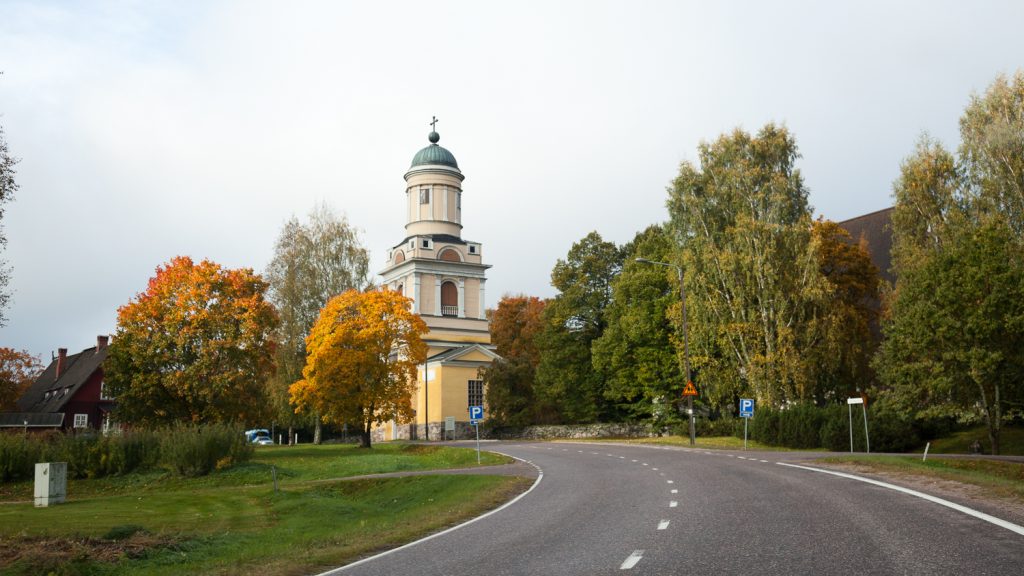 Kuvan keskellä on kellertävä kirkon kellotapuli, jonka ympärillä on lehtipuita ja edustalla tie.