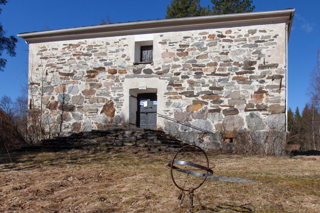 A storehouse made from natural stone, partially whitewashed, with a sun dial amidst dry grass in front of it.