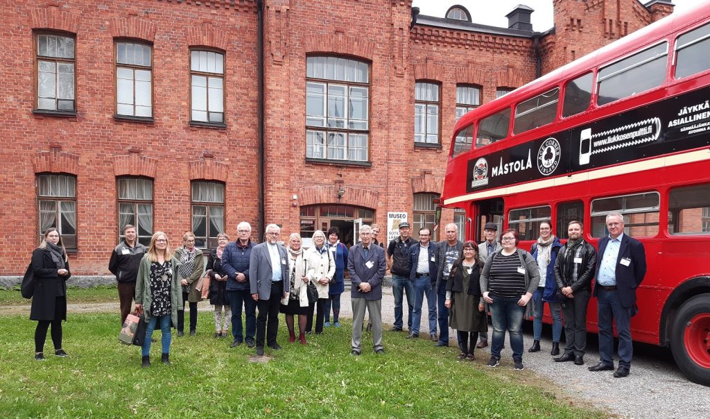 Joukko ihmisiä valokuvattavana punaisen kaksikerroksisen bussin edessä. Taustalla punatiilinen museorakennus.