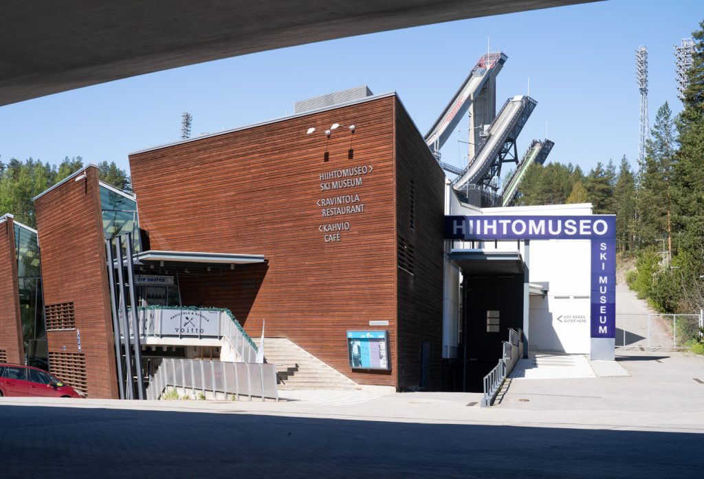 Hiihtomuseon rakennus sillan alta kuvattuna aurinkoisena päivänä. Taustalla mäkihyppytornit.