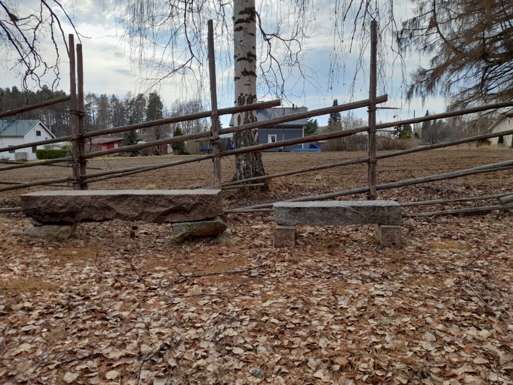 Pihakuva, nurmikolla koivunlehtiä, kaksi kivestä rakennettua penkkiä, joiden takana riukuaita, koivu ja peltoa. Taustalla taloja.